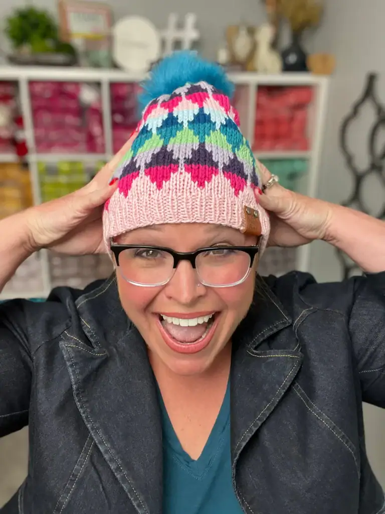 A smiling person wearing glasses and a colorful knit hat with a blue pom-pom holds the sides of the hat. Shelves filled with colorful yarn and free knitting and crochet patterns are visible in the background. -Marly Bird