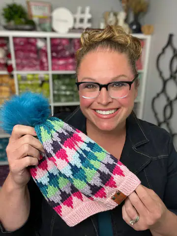 A smiling woman with glasses holds up a colorful knitted hat with a blue pom-pom, made from free knitting and crochet patterns. The diamond-patterned hat shines in pink, green, blue, purple, and white. Yarn skeins sit on shelves behind her. -Marly Bird
