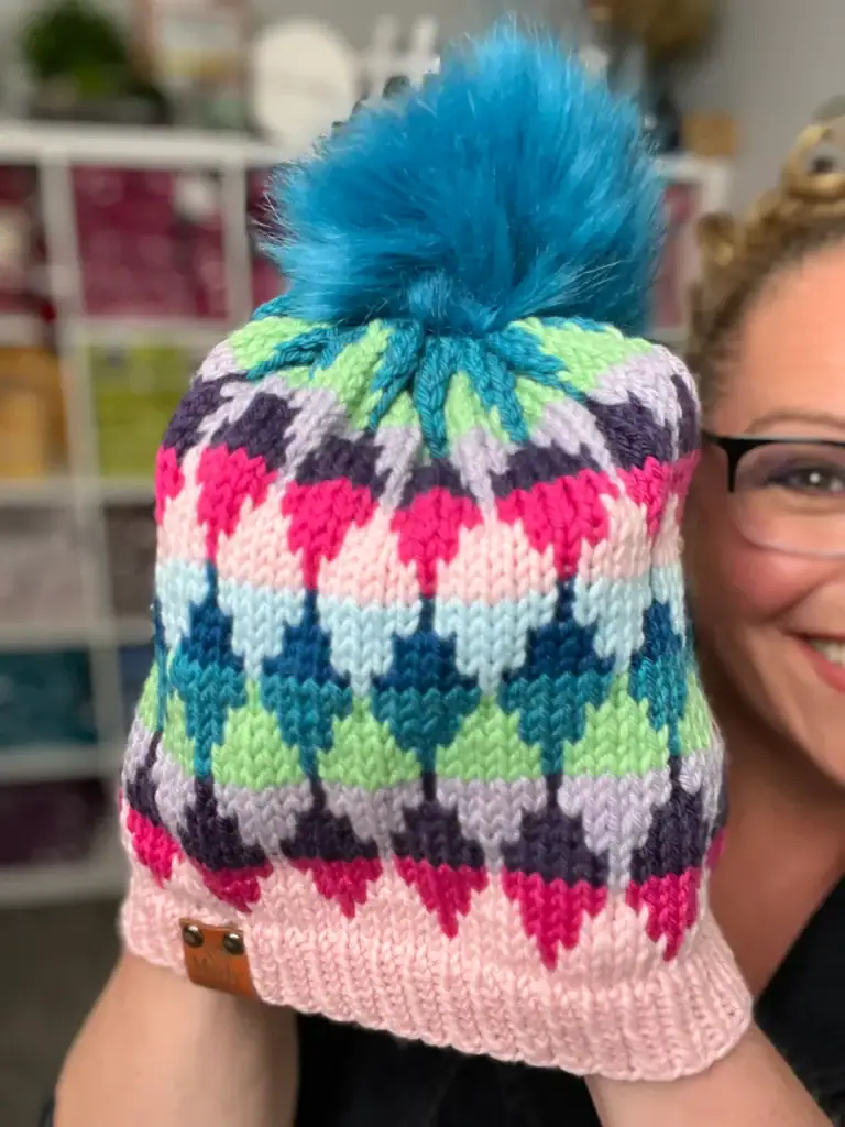 A person holds up a colorful knitted hat with a large blue pom-pom, made using free knitting and crochet patterns. The hat features a diamond pattern in pink, purple, green, and blue. The person is smiling with shelving in the background. -Marly Bird