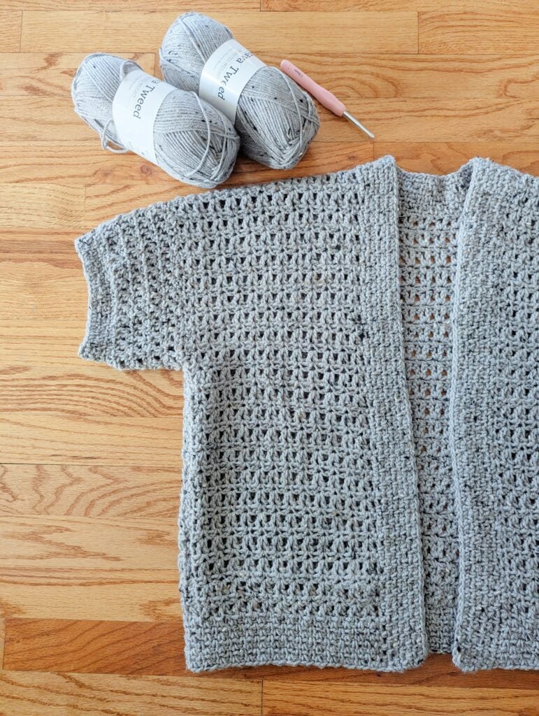 Light gray crochet cardigan with visible stitch texture, laid flat beside two gray yarn skeins and a pink-handled crochet hook.