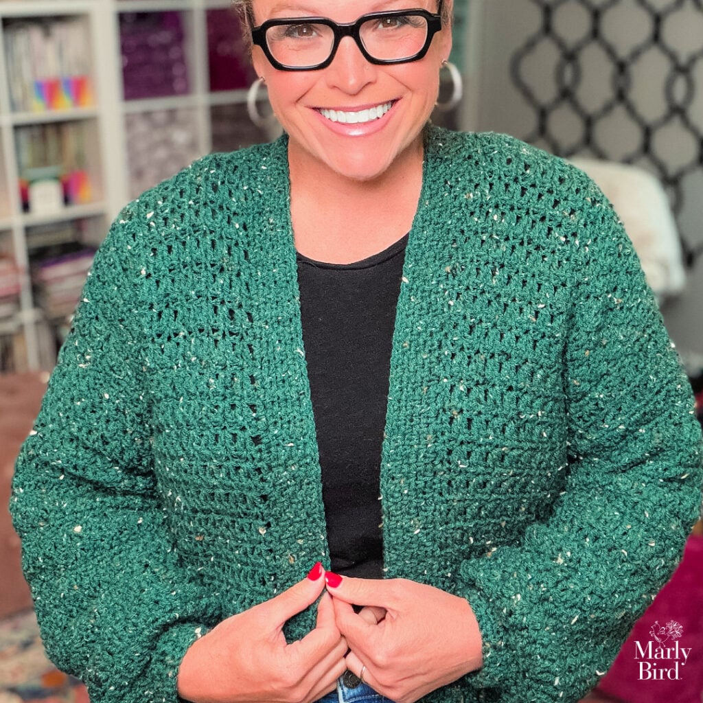 Person models a size-inclusive green crochet cardigan with rich texture and stitch detail, worn over a black shirt indoors.