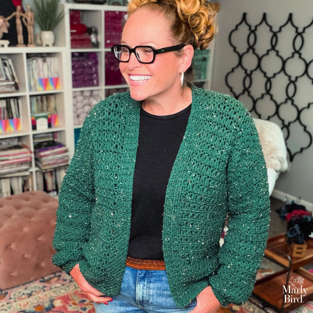A woman models a green crochet cardigan with textured stitches, shown over a black shirt and jeans in a cozy room with yarn and books.