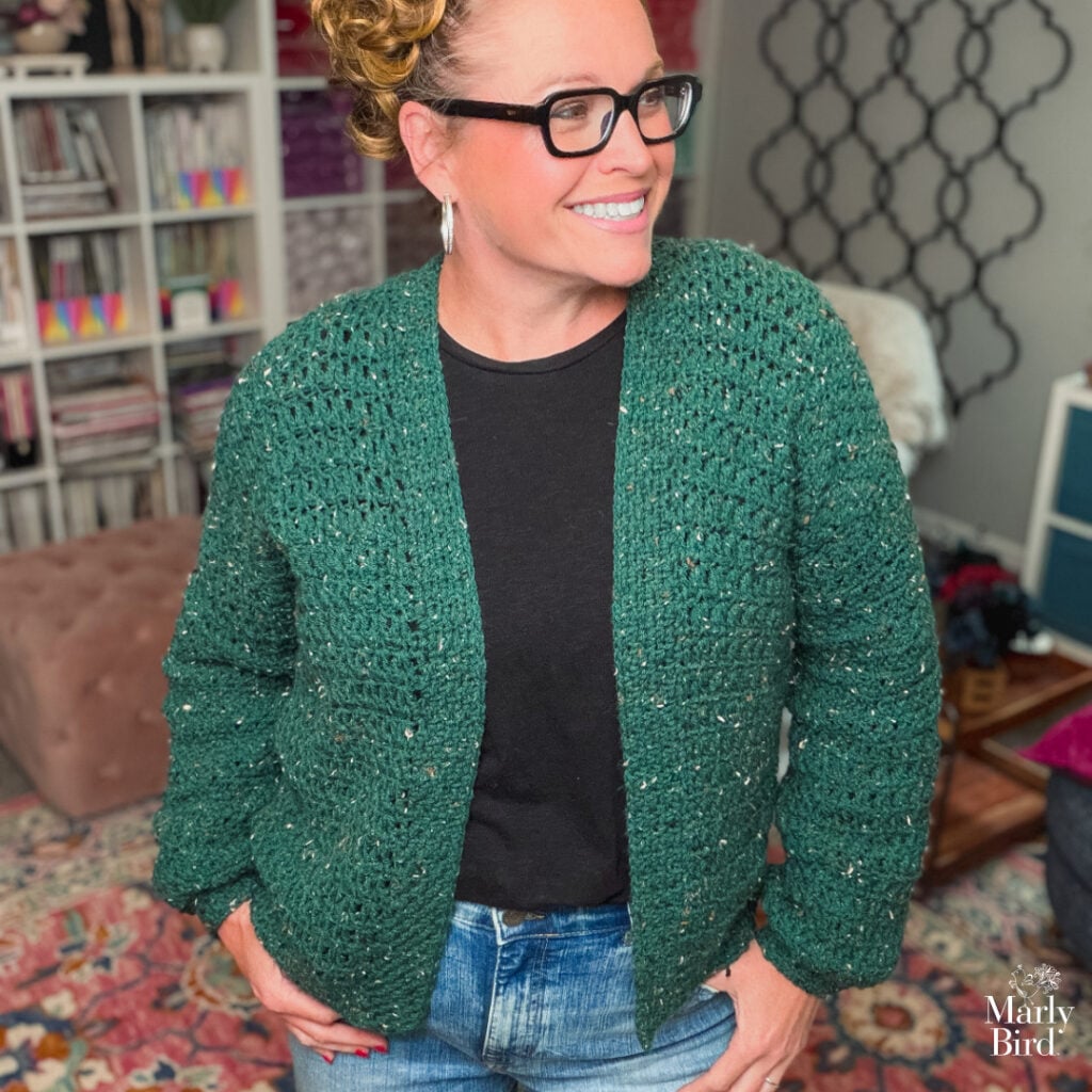 Woman models a dark green crochet cardigan with visible textured stitches in a cozy, colorful room with bookshelves.