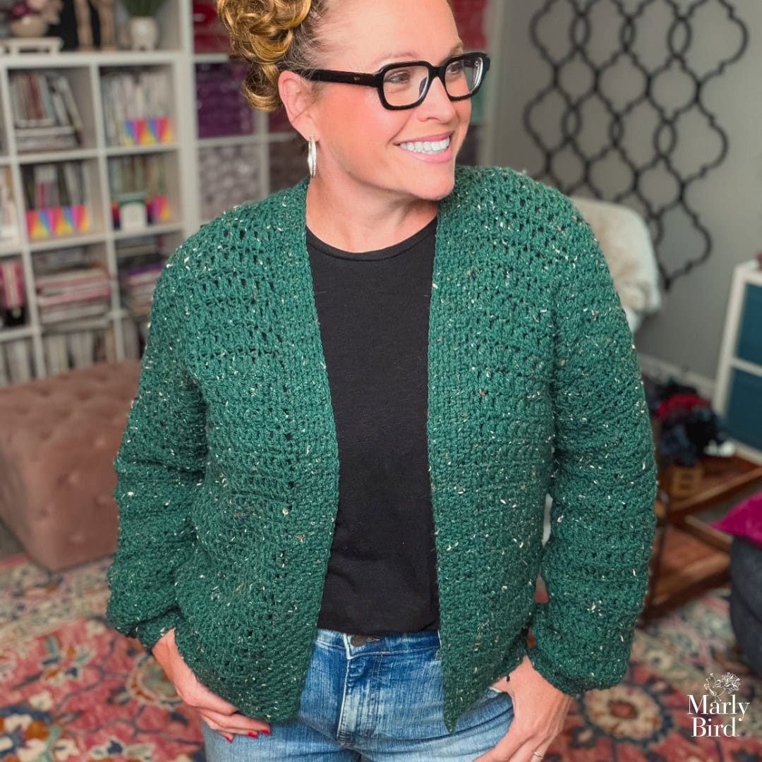 Woman models a dark green crochet cardigan with visible textured stitches in a cozy, colorful room with bookshelves.