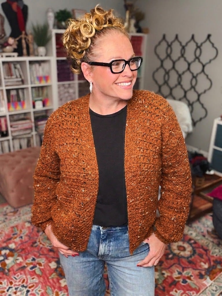 Woman models a textured orange-brown crochet cardigan, showing its stitch detail; worn over black shirt and blue jeans.