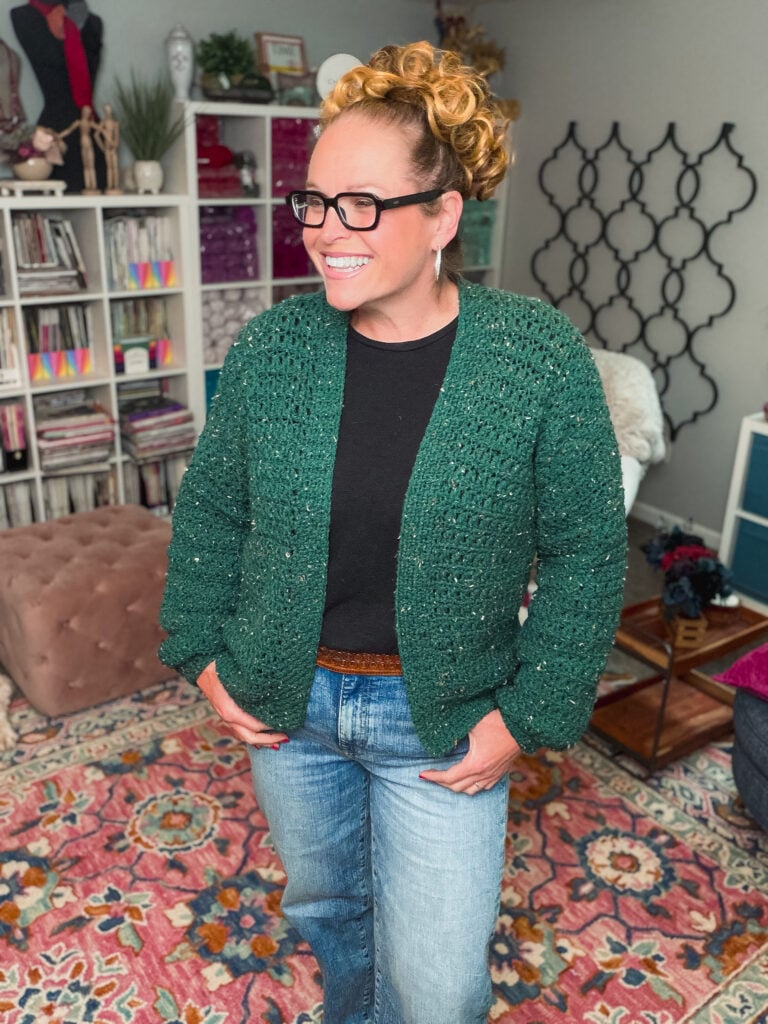 Woman models a green crochet cardi with textured stitching over a black shirt, displayed in a colorful, book-filled craft room.