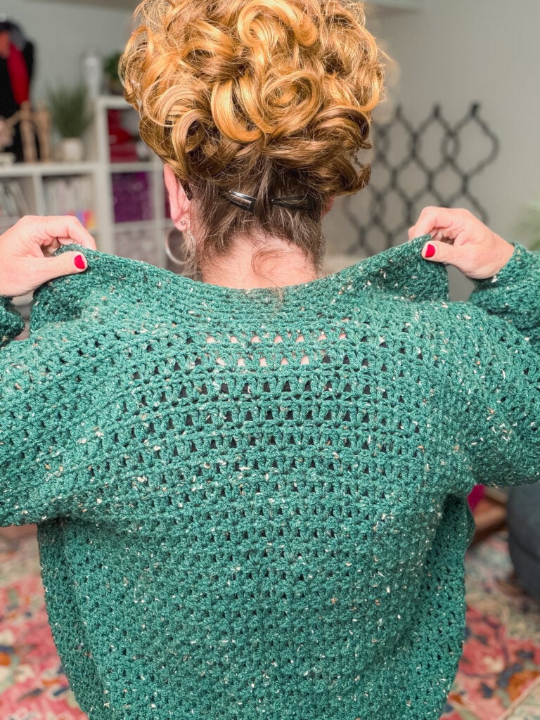 A green crochet cardigan with open stitch detail is modeled indoors, highlighting texture and size-inclusive fit.