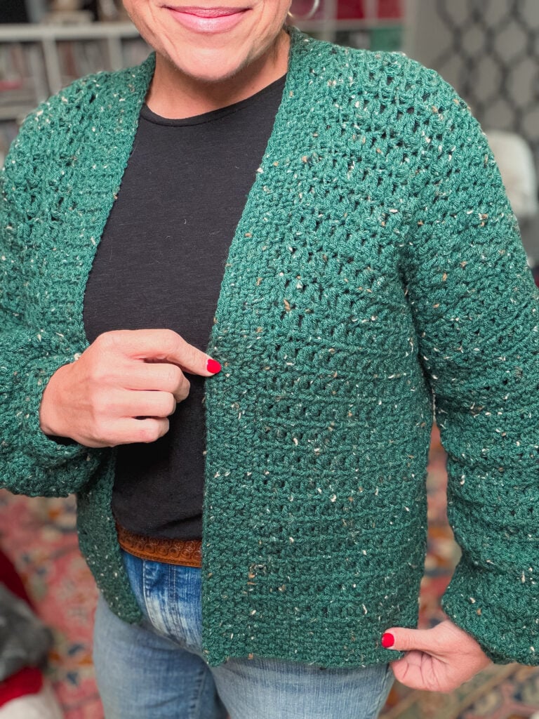 Green crochet cardigan with visible stitch texture, worn over black shirt and blue jeans; red nails and rug add contrast.