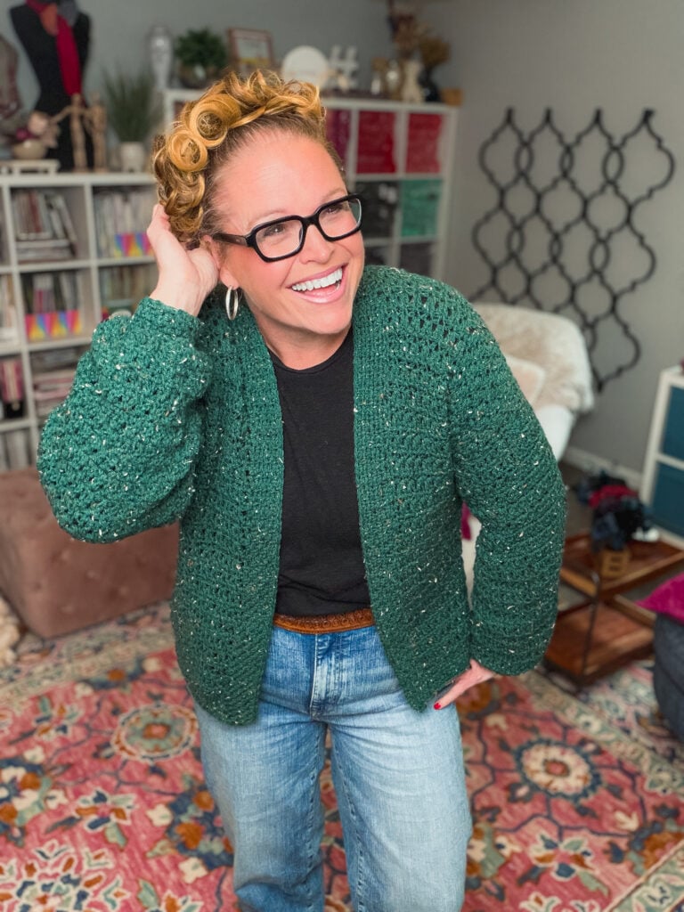 A woman models a green crocheted quick cardi with visible textured stitches, paired with a black shirt and blue jeans in a craft room.