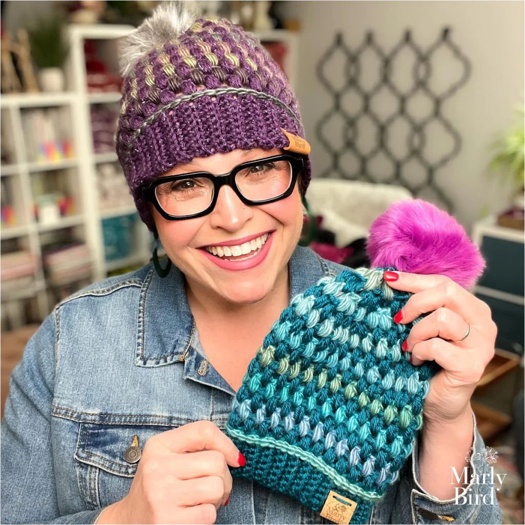 Person displays a blue and green Puff Stitch Hat with a pink pom-pom; yarn and books on shelves in the background.