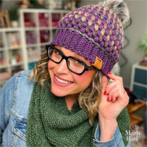 Woman models a purple and gold puff stitch crochet hat with pom-pom, paired with a green knit scarf; yarn shelves in background.