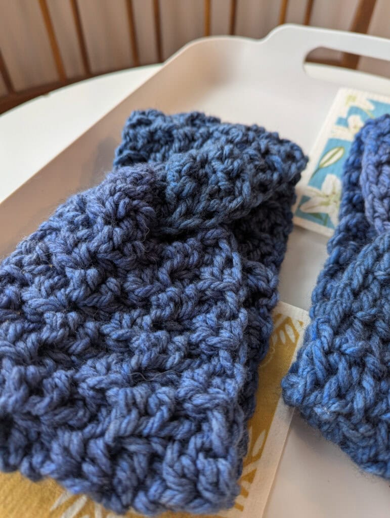 Close-up of a pair of blue crocheted fingerless gloves resting on a white tray with a partially visible floral patterned item beneath. Background shows blurred wooden chair slats. -Marly Bird