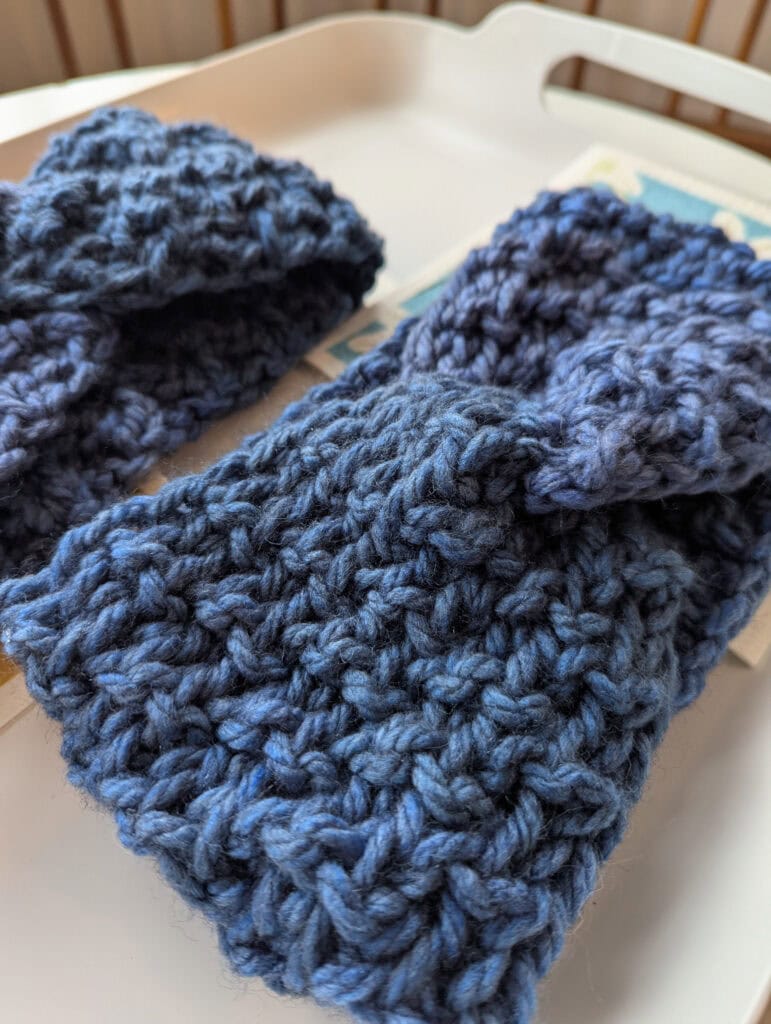 Close-up of two handmade, chunky-knit blue fingerless gloves on a white surface. The yarn has a textured, woven pattern, with varying shades of blue. The gloves are neatly placed side by side. -Marly Bird