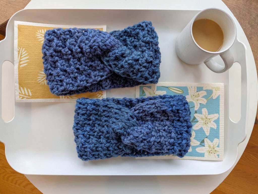 Two knitted blue headbands are placed on a white tray next to a mug of coffee. The headbands are resting on yellow and blue floral-patterned napkins. The tray is on a wooden surface. -Marly Bird