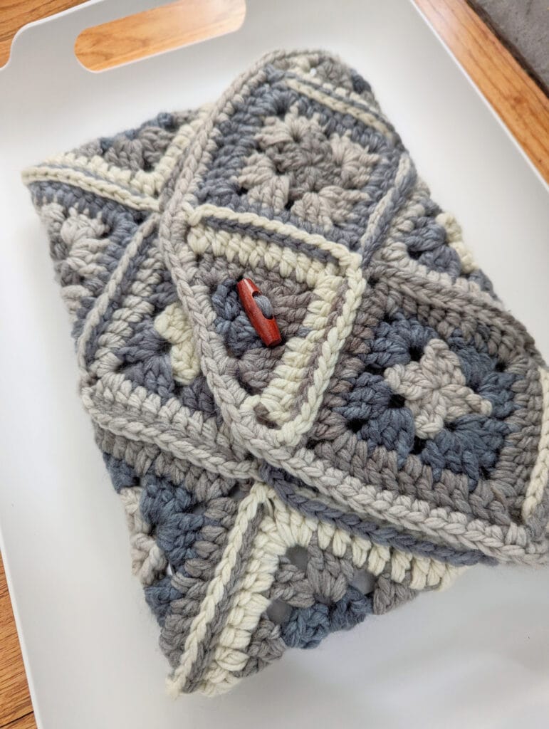 A crocheted iPad cozy, crafted from One Ball Wonders, features multi-colored granny squares in shades of gray, cream, and blue. Displayed on a white tray, the cozy is complete with a flap secured by a rustic brown button for closure. -Marly Bird