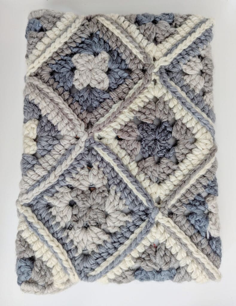 A folded crochet blanket featuring a pattern of granny square motifs in shades of grey, blue, and cream. The design includes textured stitches and a geometric arrangement. -Marly Bird