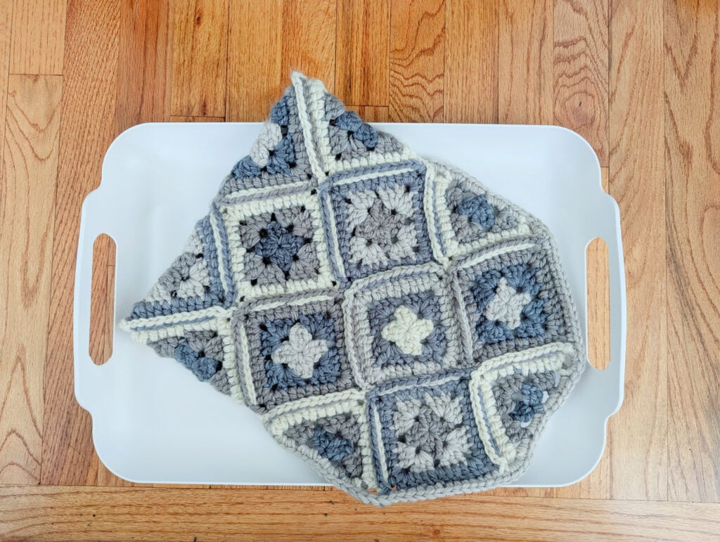 A crocheted pattern blanket featuring granny squares in shades of gray and white is folded neatly on a white tray. The tray rests elegantly on the wooden floor. -Marly Bird