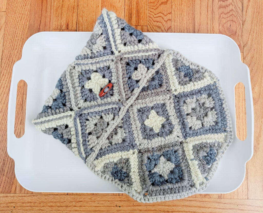 A crocheted iPad cozy featuring grey, cream, and blue granny squares with a heart-shaped button sits on a white tray with handles. The wooden floor beneath adds warmth to the scene. -Marly Bird