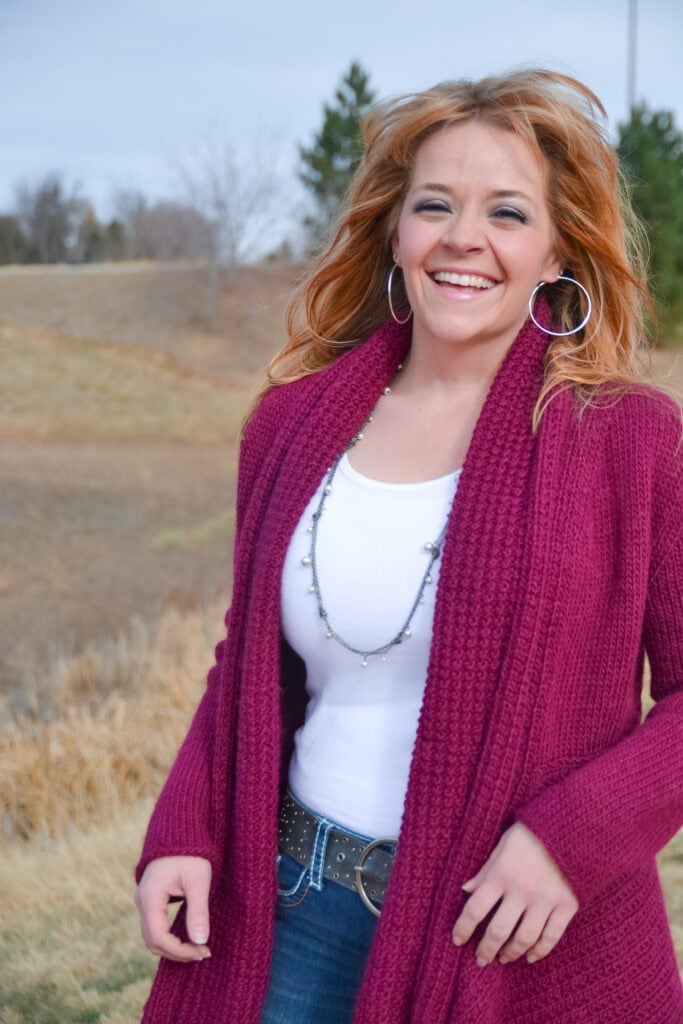 A woman with wavy red hair, wearing a white top, long maroon sweater inspired by a free knit swing cardigan pattern, and hoop earrings, smiles outdoors in a grassy, lightly wooded area on a cloudy day. -Marly Bird