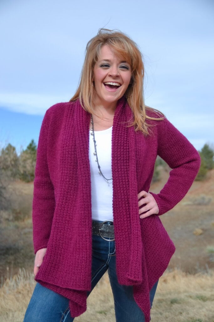A woman with blonde hair, wearing a magenta cardigan made from a free knit swing cardigan pattern, white top, and blue jeans, stands outdoors smiling with one hand on her hip against a backdrop of trees and blue sky. -Marly Bird