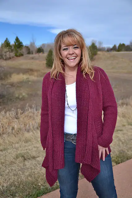 A woman stands outdoors on a path, smiling at the camera in a maroon cardigan made from free knitting and crochet patterns. She pairs it with a white top, blue jeans, and a necklace. The background features grass, trees, and a partly cloudy sky. -Marly Bird