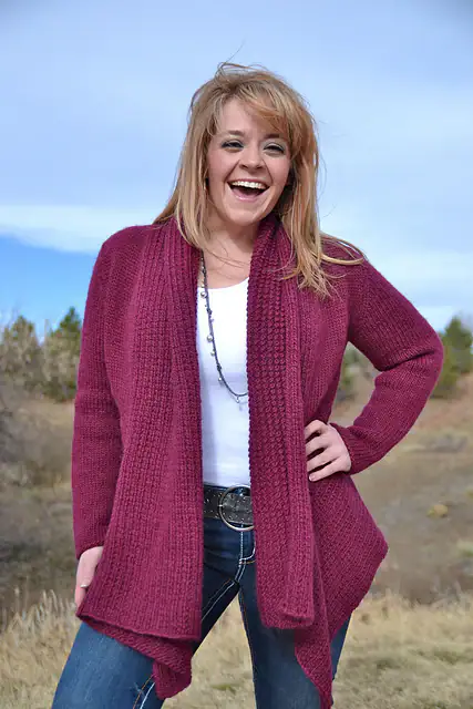 A woman stands outdoors smiling, wearing a maroon cardigan over a white top with jeans. She has light brown hair and is surrounded by trees and blue sky—her cozy look perfect for showing off free knitting and crochet patterns. -Marly Bird