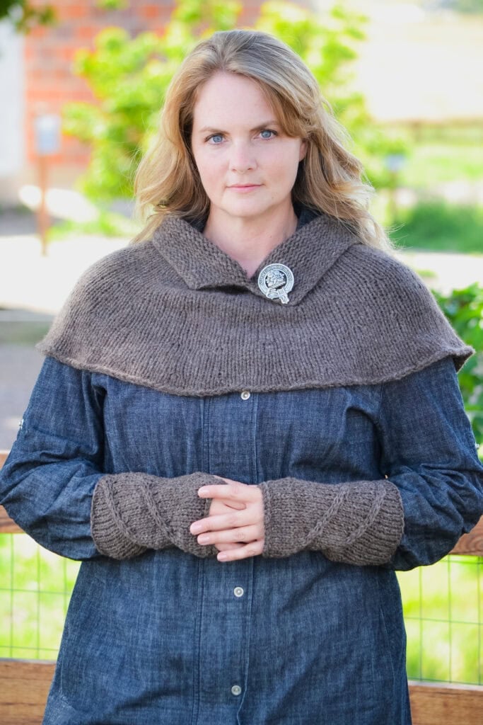 Woman modeling a brown knitted Blood of My Blood shawl with brooch and matching wrist warmers, showing stitch details.