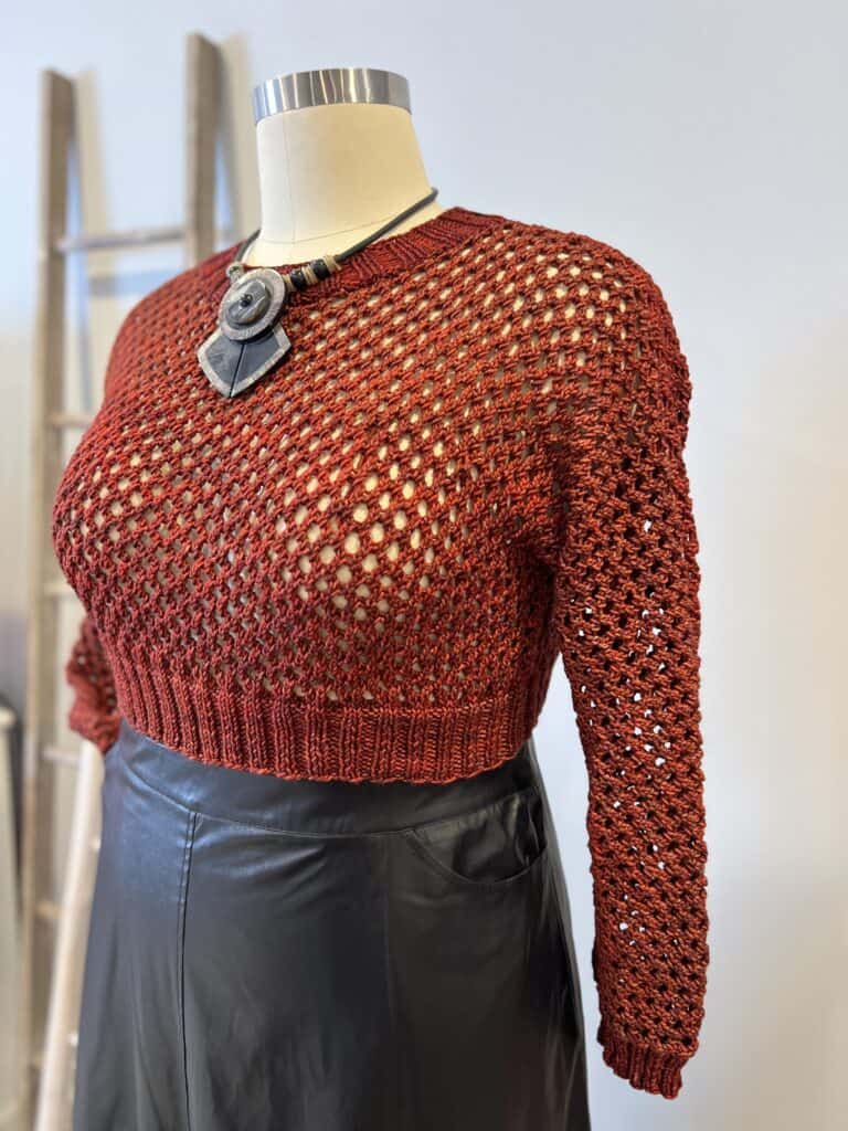 A mannequin is dressed in a rust-colored, hand-knitted, open-weave sweater paired with a black leather skirt. The outfit is accessorized with a large, round, statement necklace. A wooden ladder is visible in the background. -Marly Bird