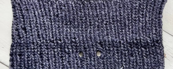 Close-up of a knitted swatch in dark gray yarn with a vertical ribbed pattern. Two small holes hint at a buttonhole feature, perfect for Game Day attire. The swatch is placed on a light-colored wood surface. -Marly Bird