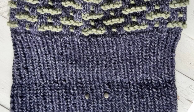 Close-up of a knitted fabric with a geometric pattern, perfect for 2025 Knit Game Day. The top showcases a textured mint green and gray diamond design, while the bottom half is a deep blue ribbed section with two small holes. The surface rests on a light wooden background. -Marly Bird