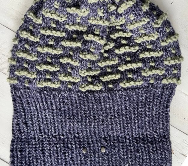 A knitted hat featuring a textured pattern with alternating blue and green yarn on the upper section is perfect for GAME DAY. The lower section is ribbed in solid blue. The hat lies flat on a wooden surface, embodying both style and team spirit. -Marly Bird