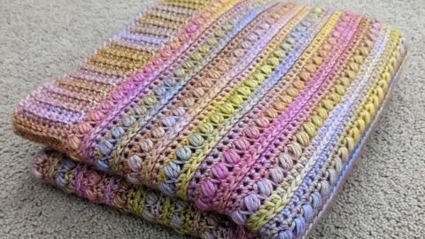 A pastel Tunisian crochet blanket, folded neatly, displays pink, purple, green, and brown stripes on a beige carpet.