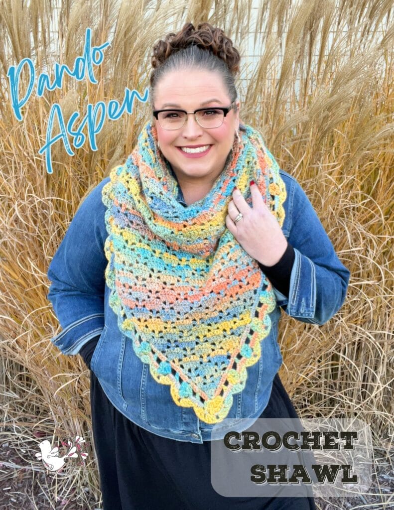 A woman wearing glasses and a colorful crochet shawl over a denim jacket stands smiling in front of tall grasses. Text on the image reads "Pando Aspen" and "Crochet Shawl." Discover Marly Bird’s crochet shawl patterns for vibrant style. -Marly Bird
