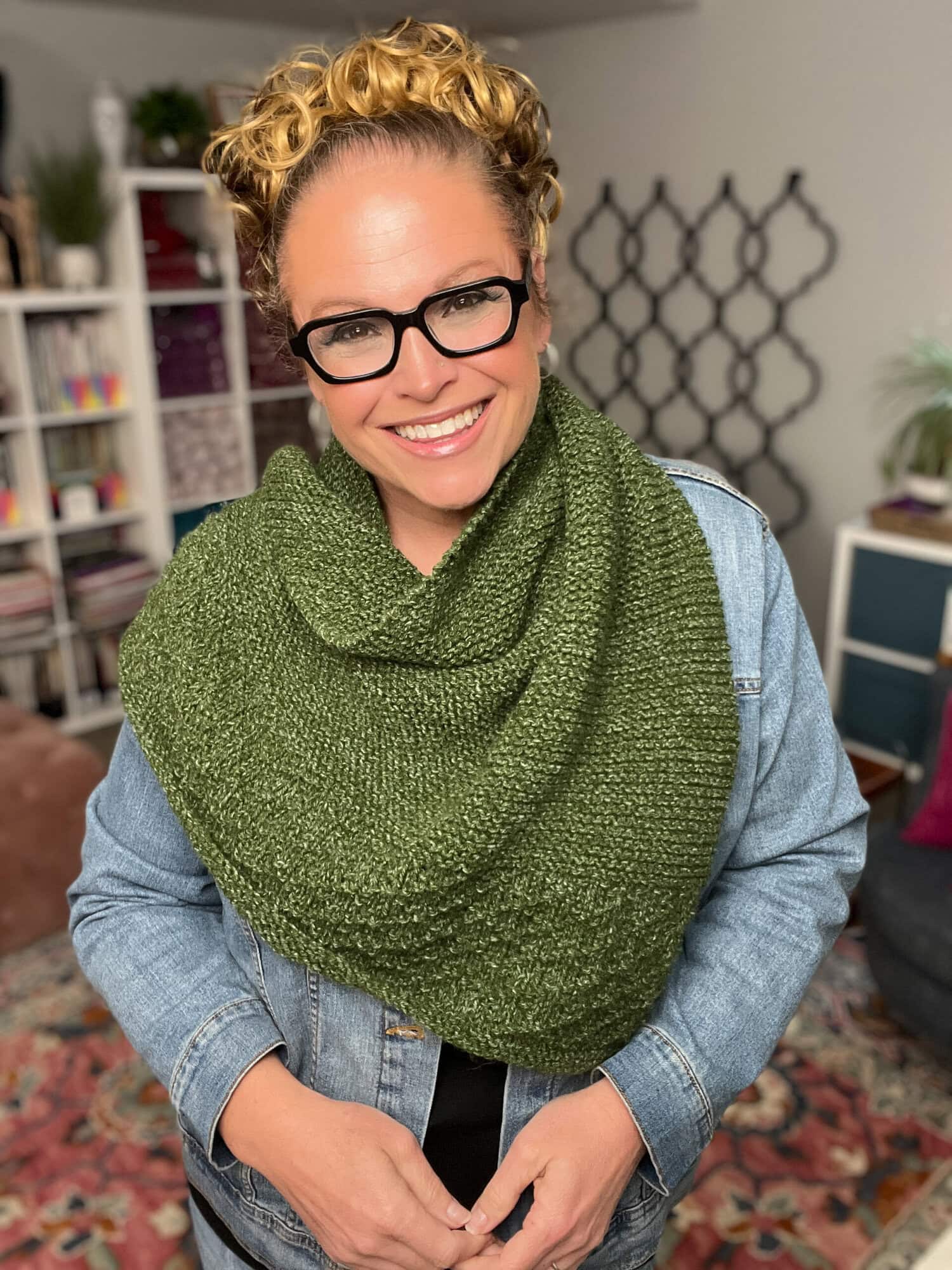 Person models a hand-knit green textured scarf indoors, with visible stitch detail; denim jacket and bookshelves behind.