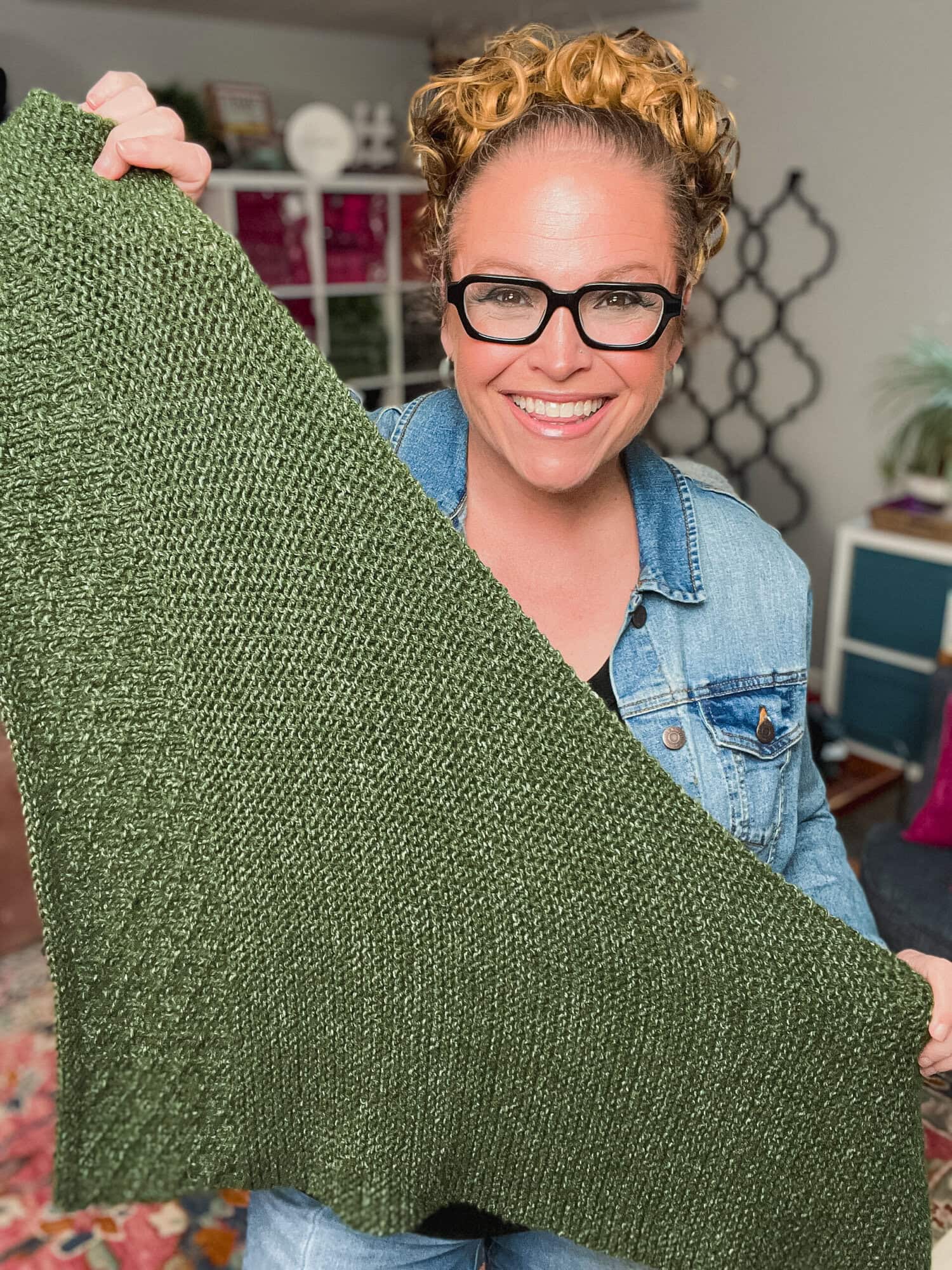Woman displays a green knit square shawl, showing off its textured stitches; yarn shelves fill the background.