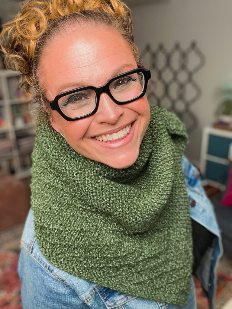 Woman models a chunky green knit shawl with textured stitches, styled over a denim jacket in a cozy, book-filled room.