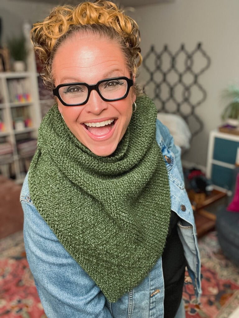 Person models a green knit scarf with textured stitches, inspired by a one ball shawl pattern, paired with a denim jacket.