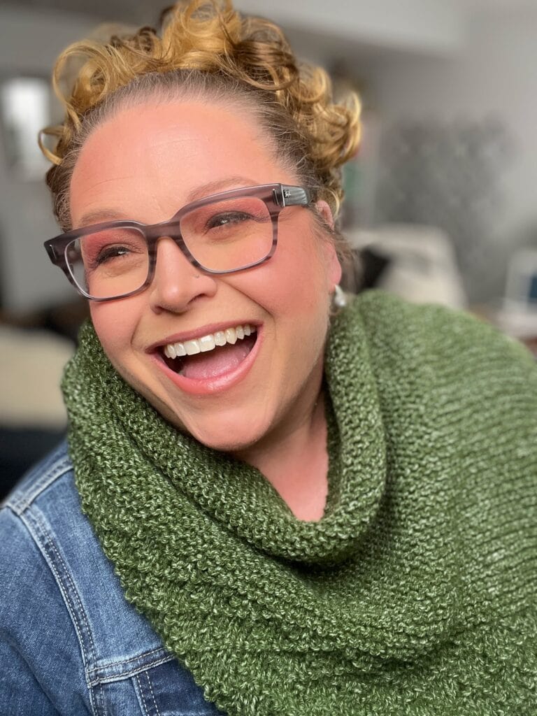 A smiling woman with curly hair and glasses, wearing a green Serenity Shawl with a denim jacket, in a relaxed setting. She exudes happiness and joy. -Marly Bird