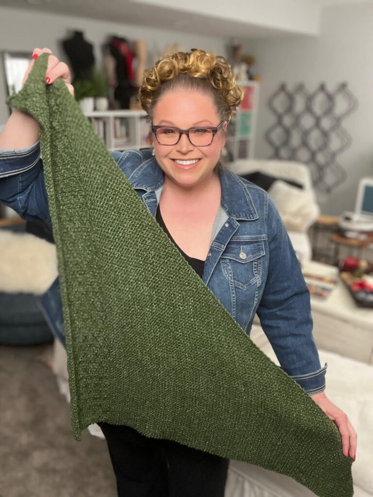 A person with curly hair and glasses smiles while holding up a green knitted triangular piece, a perfect example of Serenity Knit. They are wearing a denim jacket. The background shows a cozy room with bookshelves and patterned wall decor. -Marly Bird