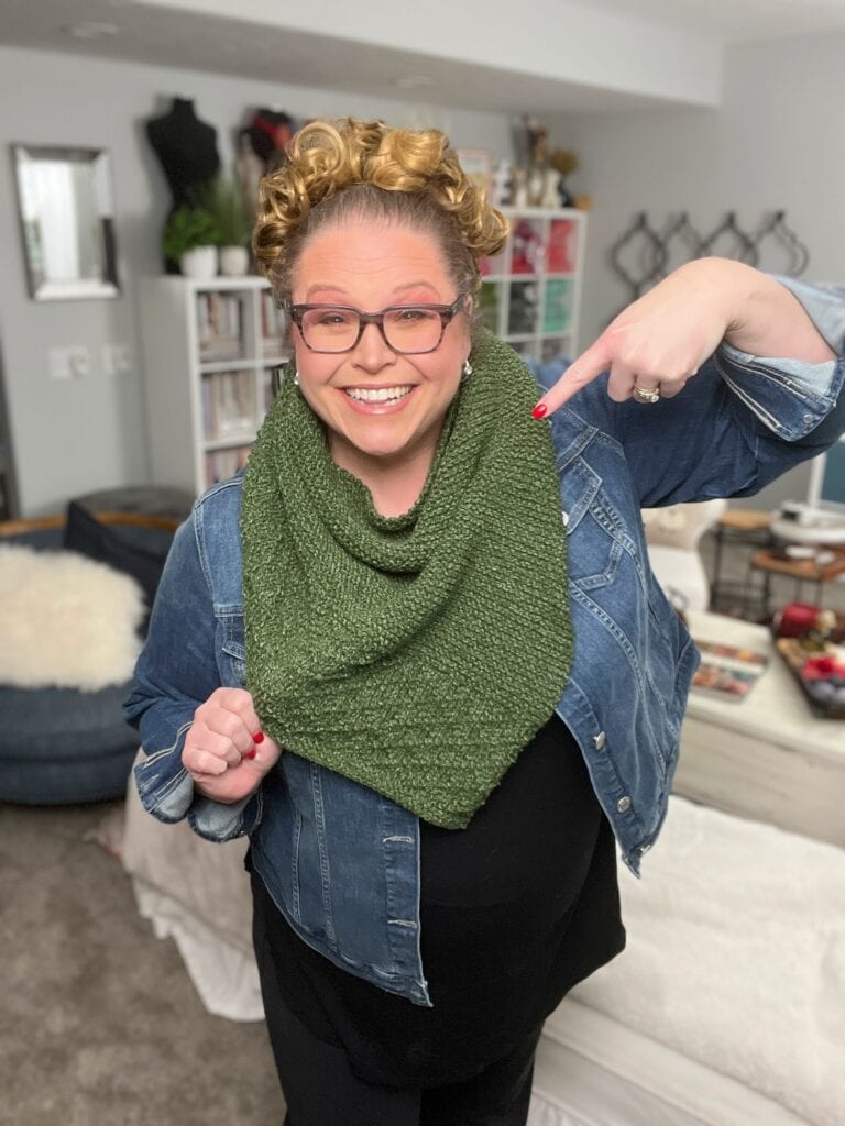 A smiling woman wearing glasses points to a green knit scarf she's fashioned from a one ball knit shawl pattern. Her hair is styled in curls, and she's dressed in a denim jacket. Behind her, a living room is visible with shelves, a white couch, and decorative items. -Marly Bird