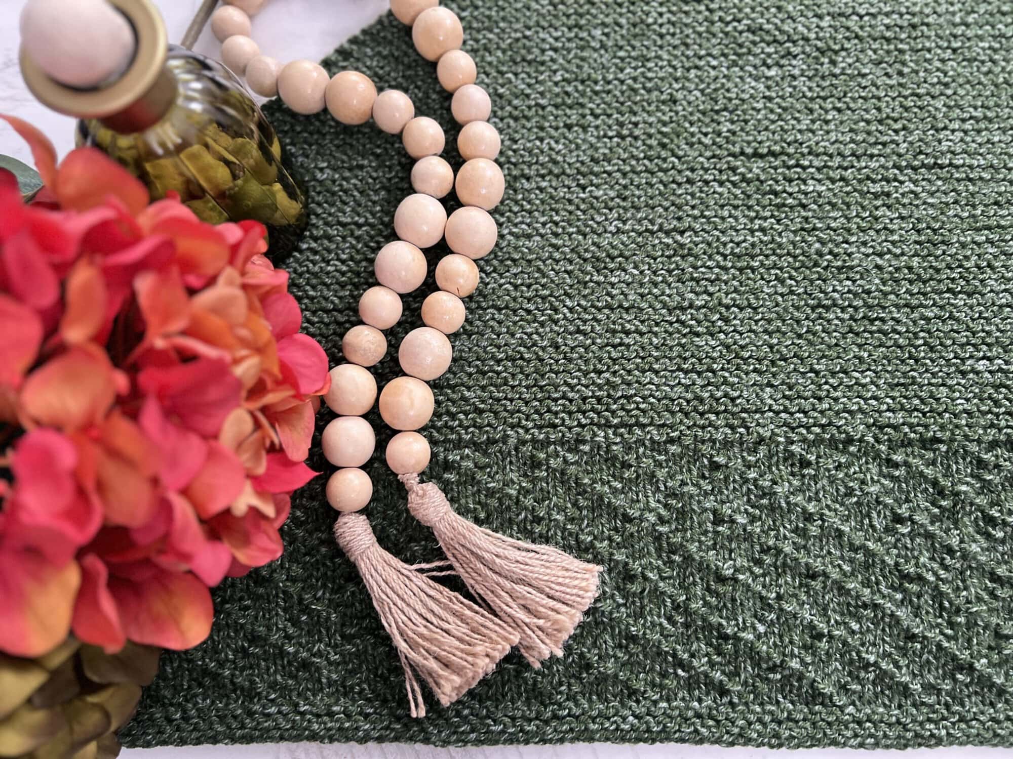 A textured green knit shawl covers most of the image. On top, a wooden bead garland with tassels is arranged beside a bouquet of red and pink hydrangeas in a vase, creating an ambiance of peak serenity. -Marly Bird