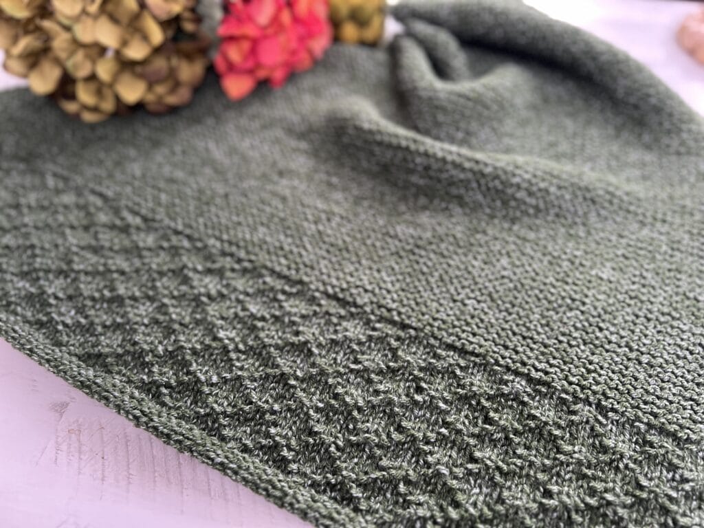 The "Serenity Knit" hat showcases a close-up of its green textured patterning on the brim. Laid on a white surface, it is accompanied by dried flowers in shades of yellow and pink, evoking a sense of calmness and beauty. -Marly Bird