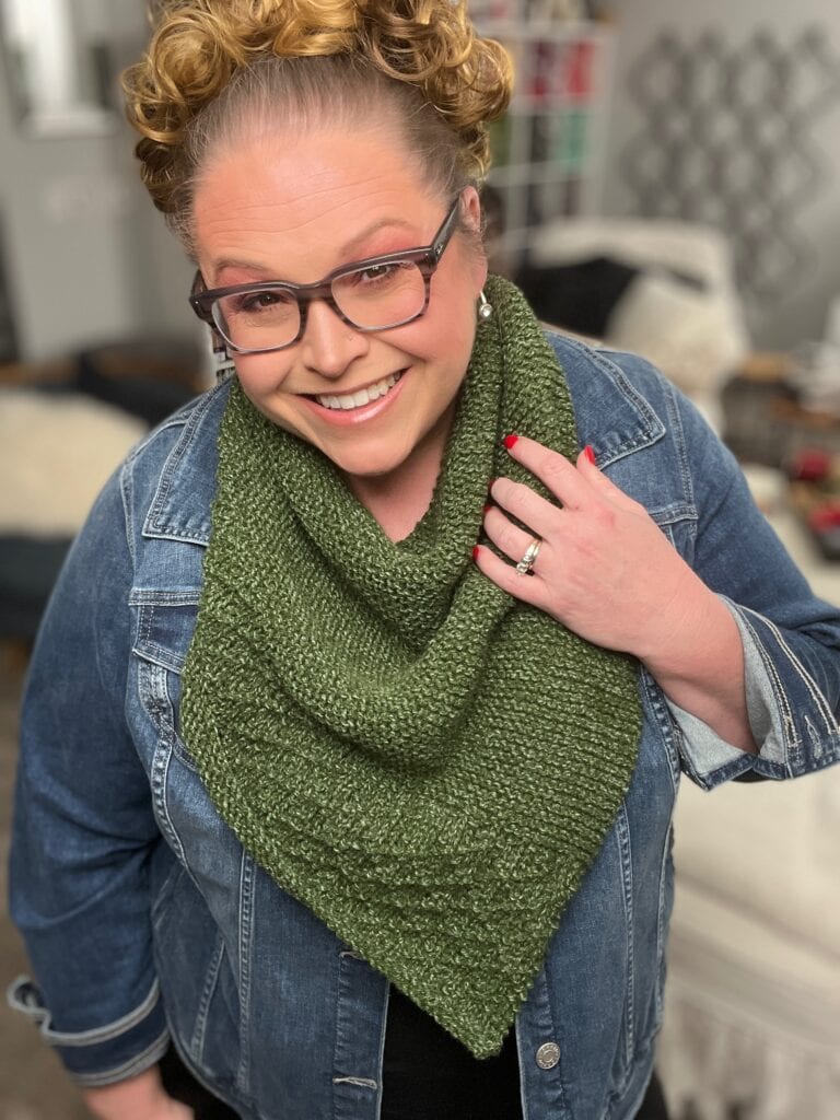 A smiling person with styled curly hair, wearing glasses, a denim jacket, and a cozy Serenity Knit Shawl. They are indoors, and the background shows a warm and inviting living room setting. -Marly Bird