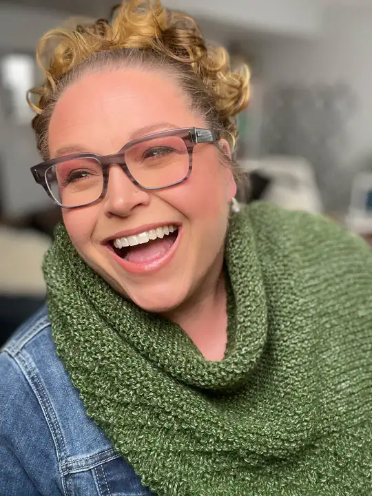 A woman with curly hair, glasses, and pink eye makeup smiles broadly. She wears a green knitted scarf—possibly inspired by free knitting and crochet patterns—and a denim jacket, with a blurred indoor background. -Marly Bird