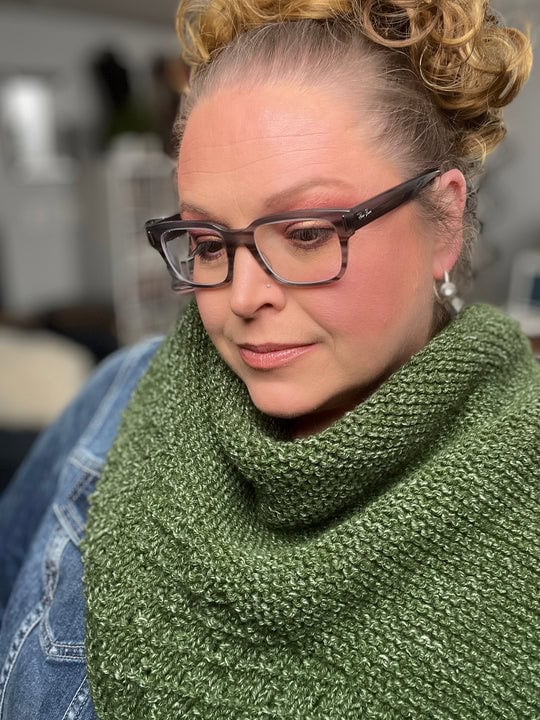 A woman with curly hair and glasses, wearing a green knit cowl from free knitting and crochet patterns and a denim jacket, looks down thoughtfully against a softly blurred indoor background. -Marly Bird