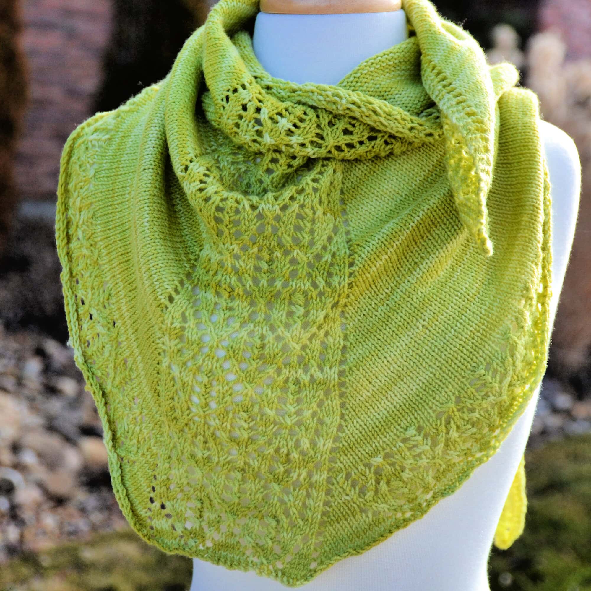 Light green knitted shawl with lace on one side and edge, displayed on a mannequin; blurred natural background.