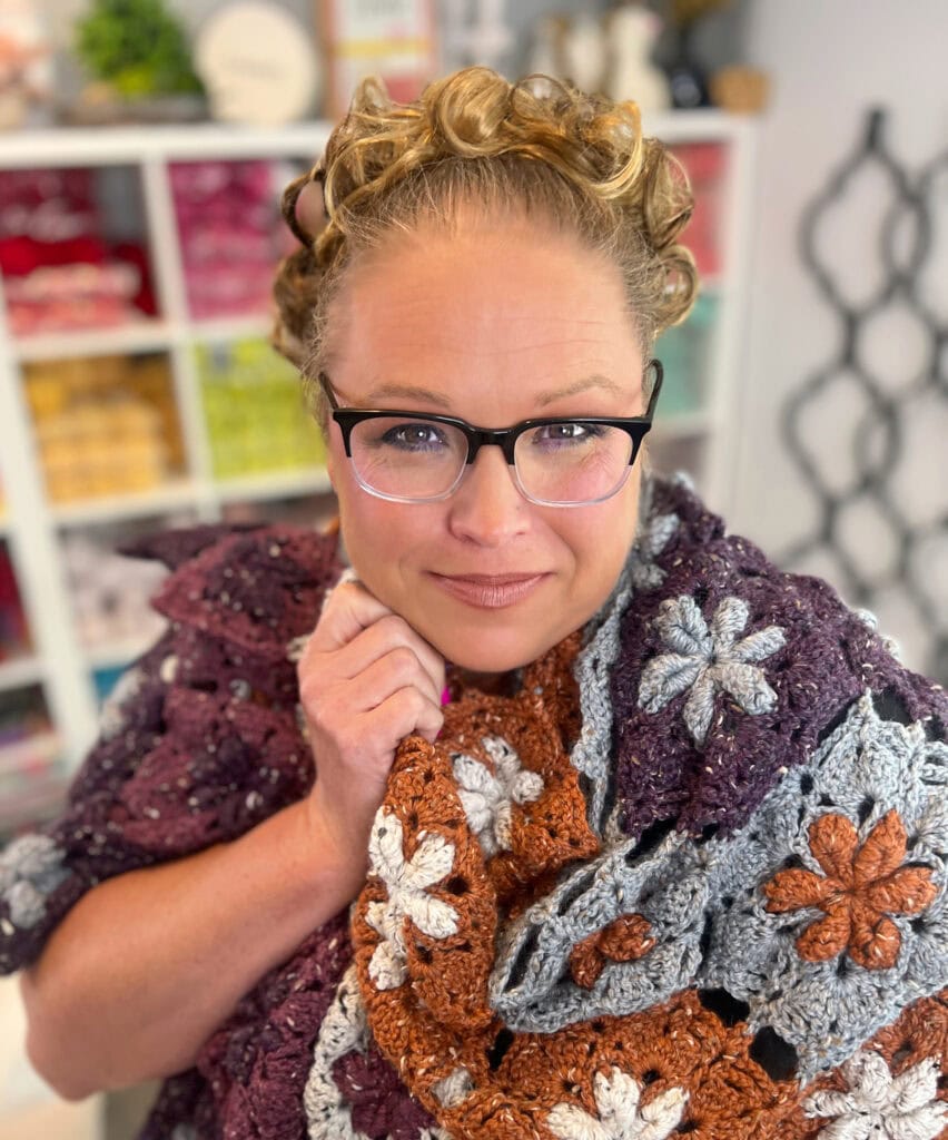 A woman with curly hair and glasses smiles softly while wrapped in a crocheted blanket with orange, gray, and white floral patterns, inspired by Marly Bird crochet shawl patterns. Shelves with colorful yarn and craft supplies are visible in the background. -Marly Bird