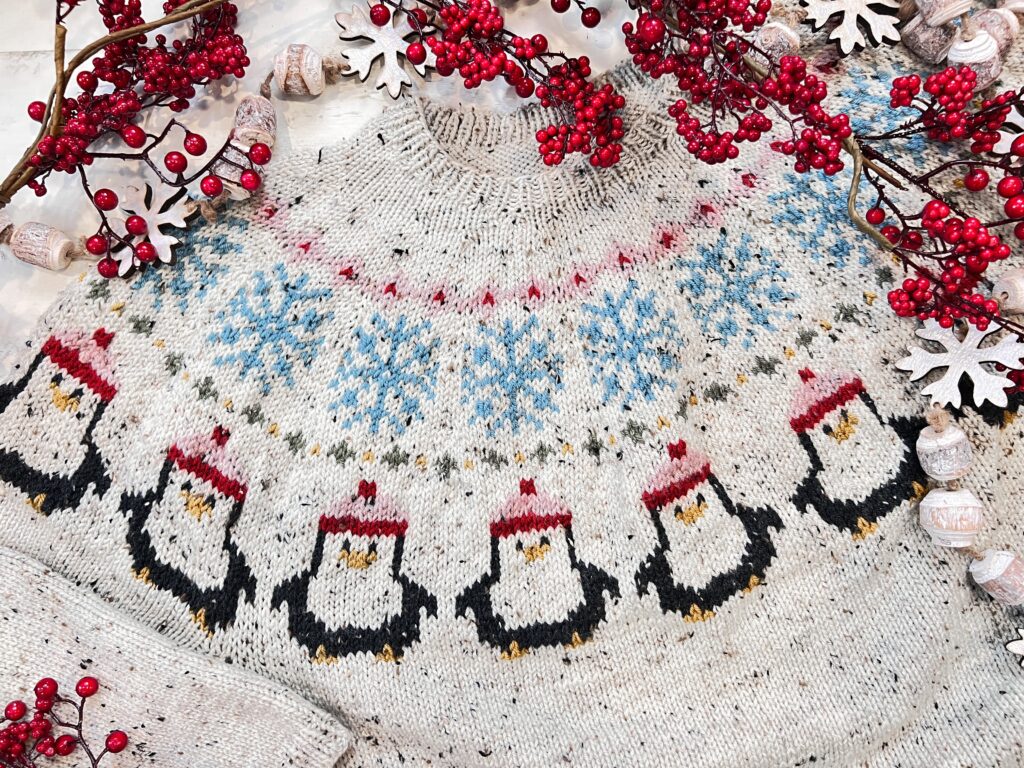A penguin yoke sweater knitting pattern featuring rows of penguins in red Santa hats, blue snowflakes, and small berry and snowflake decorations for a festive look. -Marly Bird