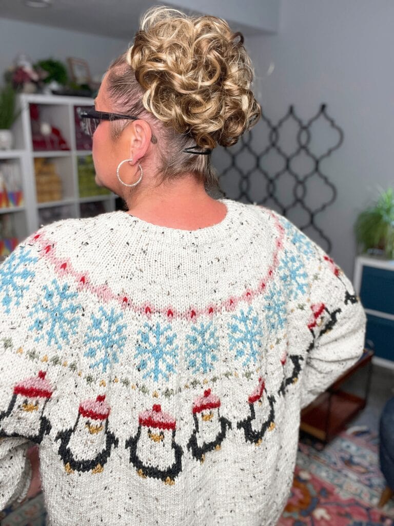 A person with curly blonde hair pulled up, wearing glasses and hoop earrings, is seen from behind in a room. They sport a white penguin yoke sweater knitting pattern featuring penguins in Santa hats and blue snowflakes. -Marly Bird