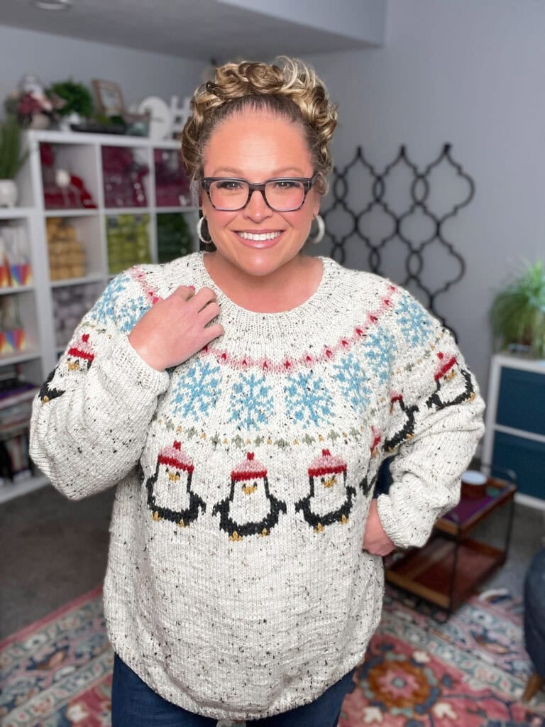 A smiling person with glasses and curly hair wears a white penguin yoke sweater knitting pattern, standing in a cozy, colorful room with shelves, plants, and decorative items. -Marly Bird